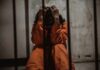 Person in orange jumpsuit sitting behind bars, handcuffed.