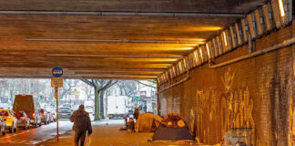 People walk under a bridge with homeless encampment