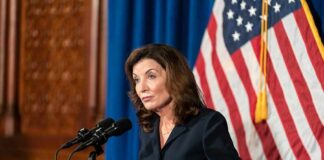 Kathy Hochul speaking at a podium during a press conference with an American flag in the background