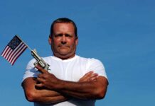 Man holding a gun and an American flag against a blue sky