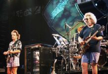 Two musicians performing on stage with guitars and a drum set in the background