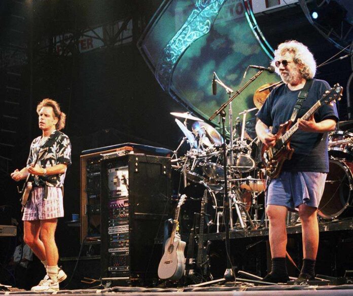 Two musicians performing on stage with guitars and a drum set in the background