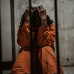 Person in orange jumpsuit sitting behind bars, handcuffed.