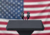 Podium with microphones, American flag background.