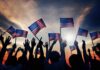 Joy Reid Torches July 4th – Backlash Erupts! Silhouetted crowd holding American flags against a sunset sky