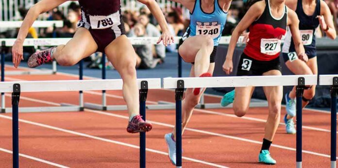 shutterstock_1970486681 Female athletes clearing hurdles on a track
