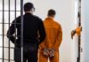Man With Mutilated Genitals ARRESTED Entering Country Illegally Prisoner in orange jumpsuit handcuffed and escorted by officer