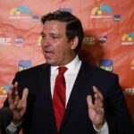 DeSantis Sounds Alarm: Institutions On The Chopping Block Man in a suit speaking at a media backdrop