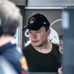 Elon Musk wearing a black cap in a crowded indoor setting