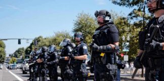 Bear Pascoe, Brian Folkerts, and Eugene Sims among riot police standing in a line on a city street