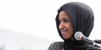 Ilhan Omar speaking at a microphone with hand on chest