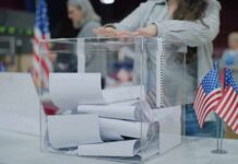 Iran Infiltrate American Voters – Dictating Midterms! Person casting a ballot into a clear box at a polling station